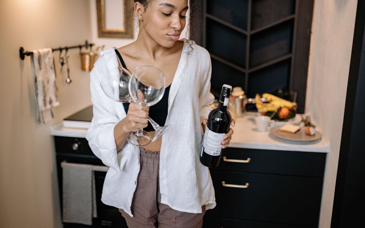 A woman with curly hair holds a bottle of wine and glasses in a stylish kitchen setting.