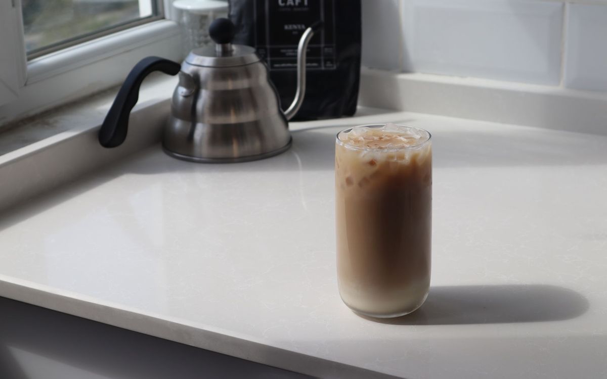 Glass of iced latte and a metal coffee kettle on a kitchen table.