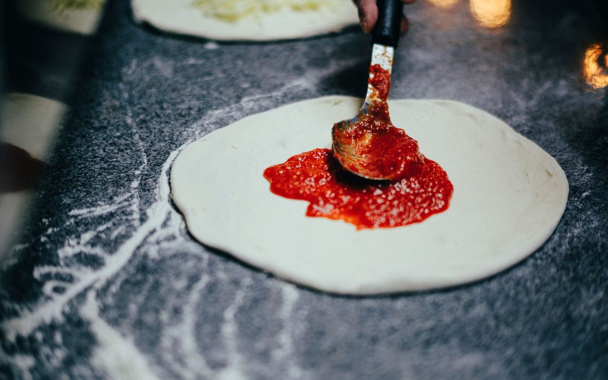 A spoon spreads tomato sauce onto a round of pizza dough on a floured counter.