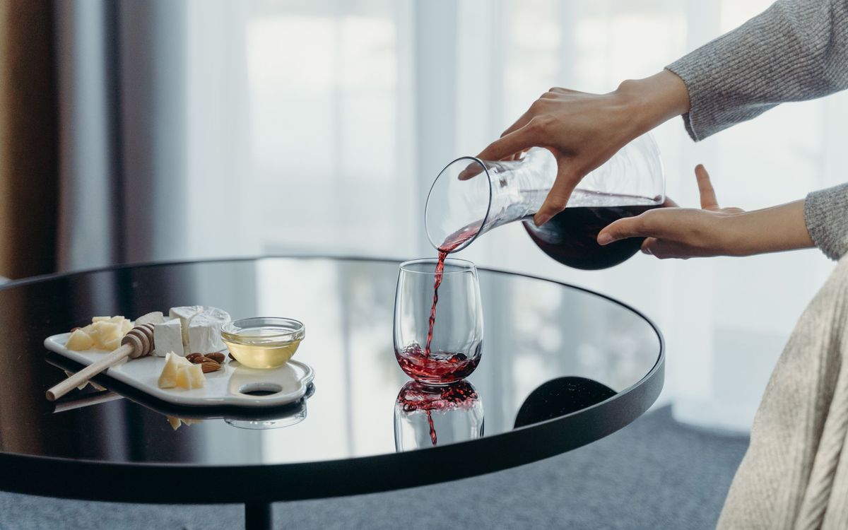 A person pours red wine into a glass next to a cheese platter on a modern glass table.