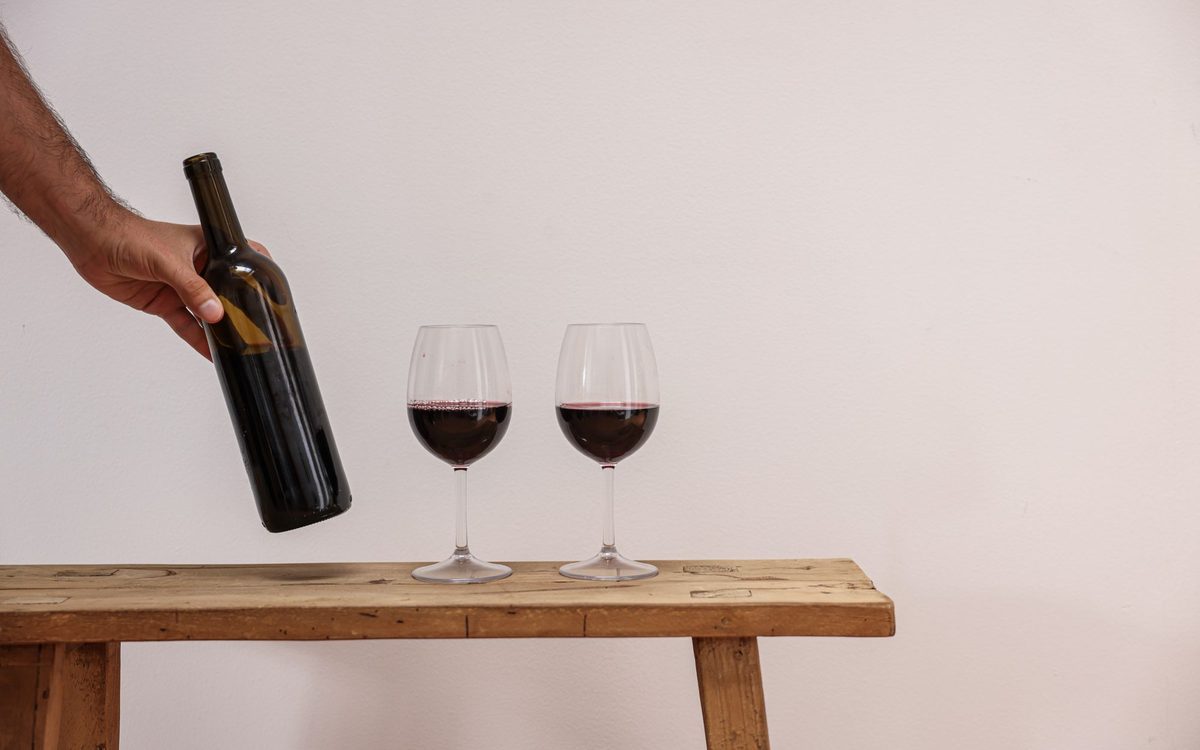 Close-up of a hand pouring red wine into two elegant glasses on a rustic wooden bench