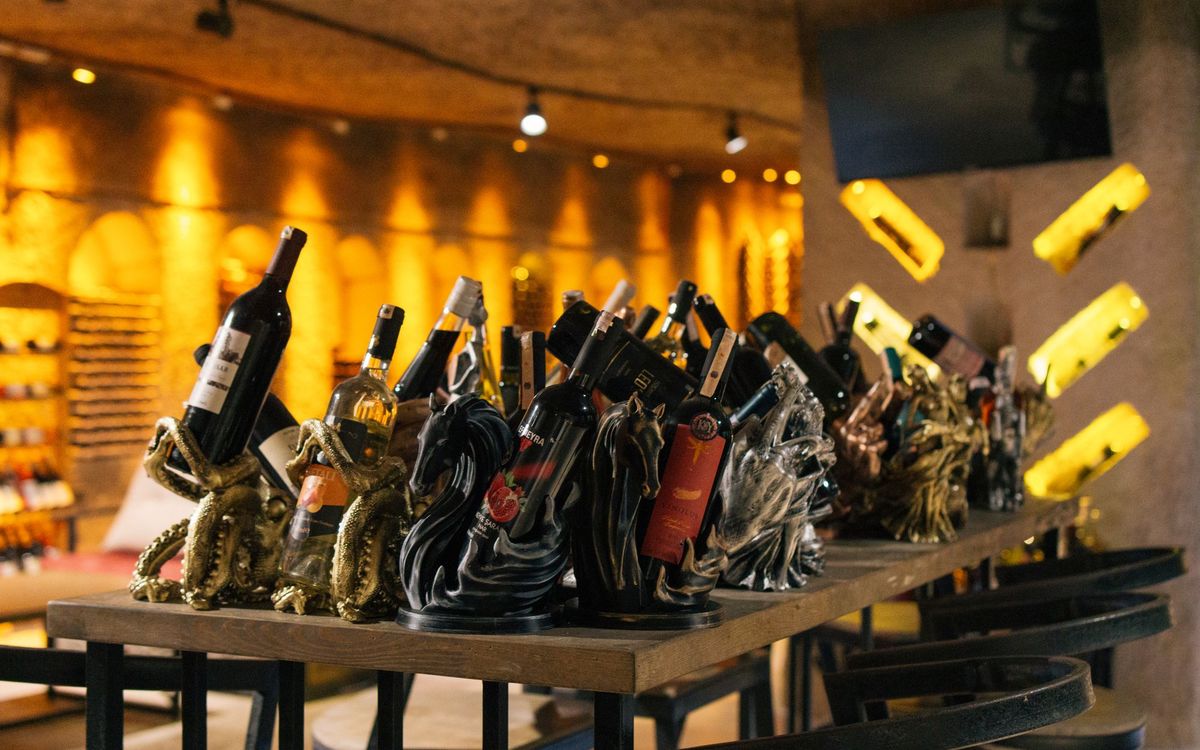 Wine bottles standing in decorative holders on a wooden bar table in a warmly lit bar, with lit wine shelves behind.