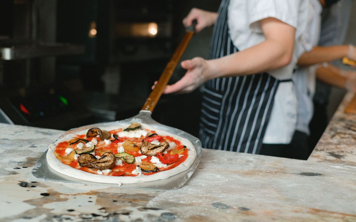 Cook sliding a topped pizza on a metal peel at a counter beside a pizzeria oven