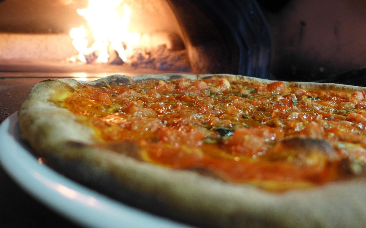 A sauced pizza on a white plate in front of a glowing wood fire inside a brick oven