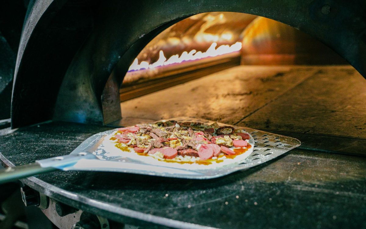 A topped pizza on a metal peel being slid onto the dark stone floor of a gas-fired pizza oven