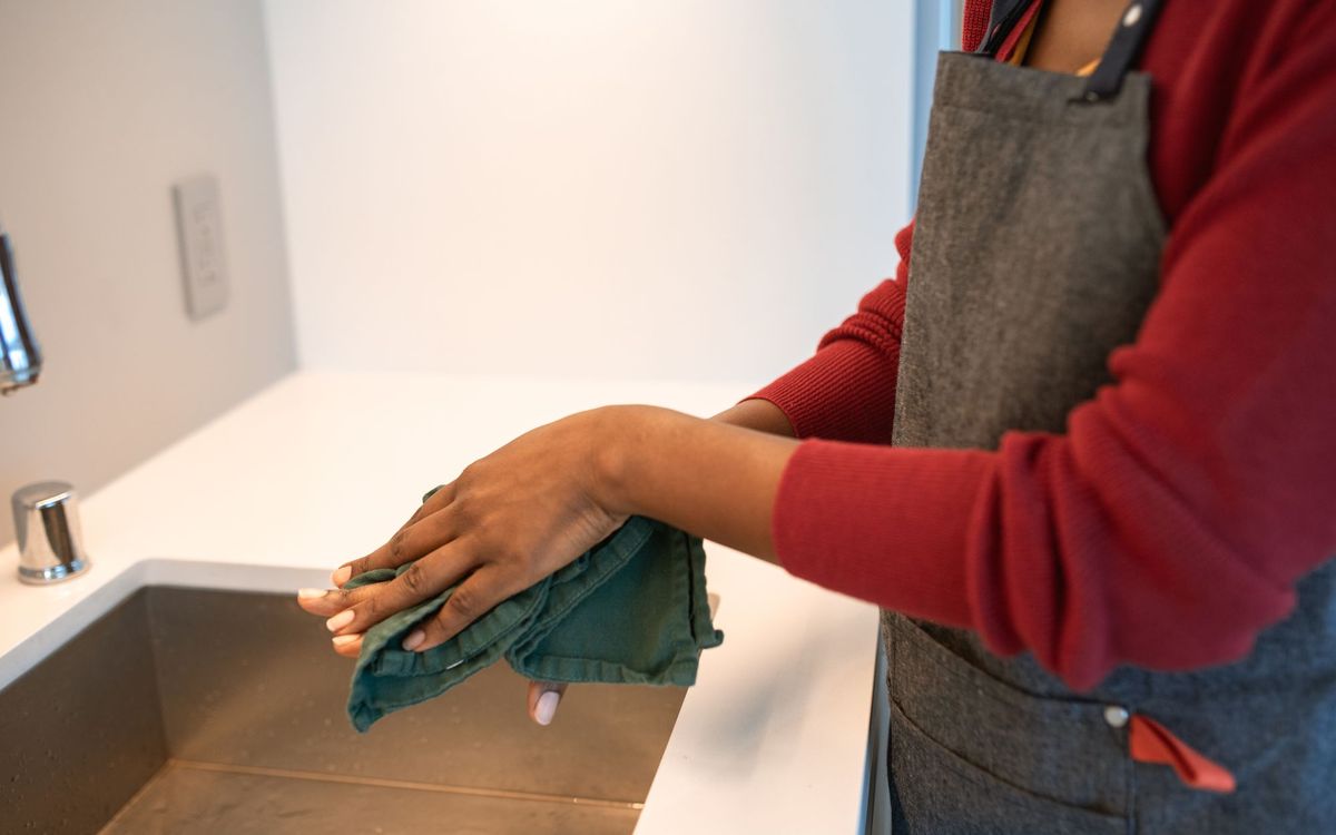Person in an apron drying their hands with a green cloth beside a kitchen sink