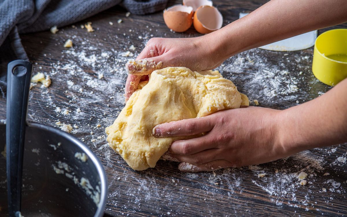 Hands pressing a yellow, egg-rich dough on a floured dark wooden table, with cracked eggshells and a measuring cup nearby.
