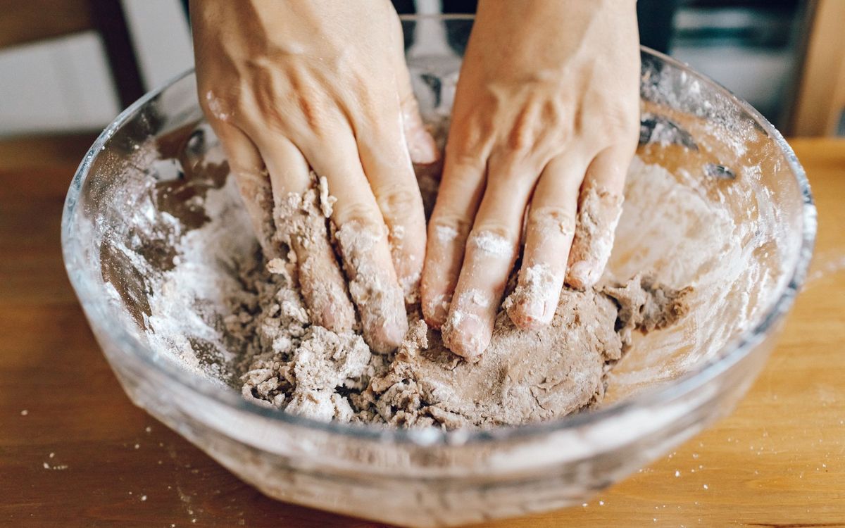 Hands working a shaggy whole grain dough in a glass bowl on a wooden table.