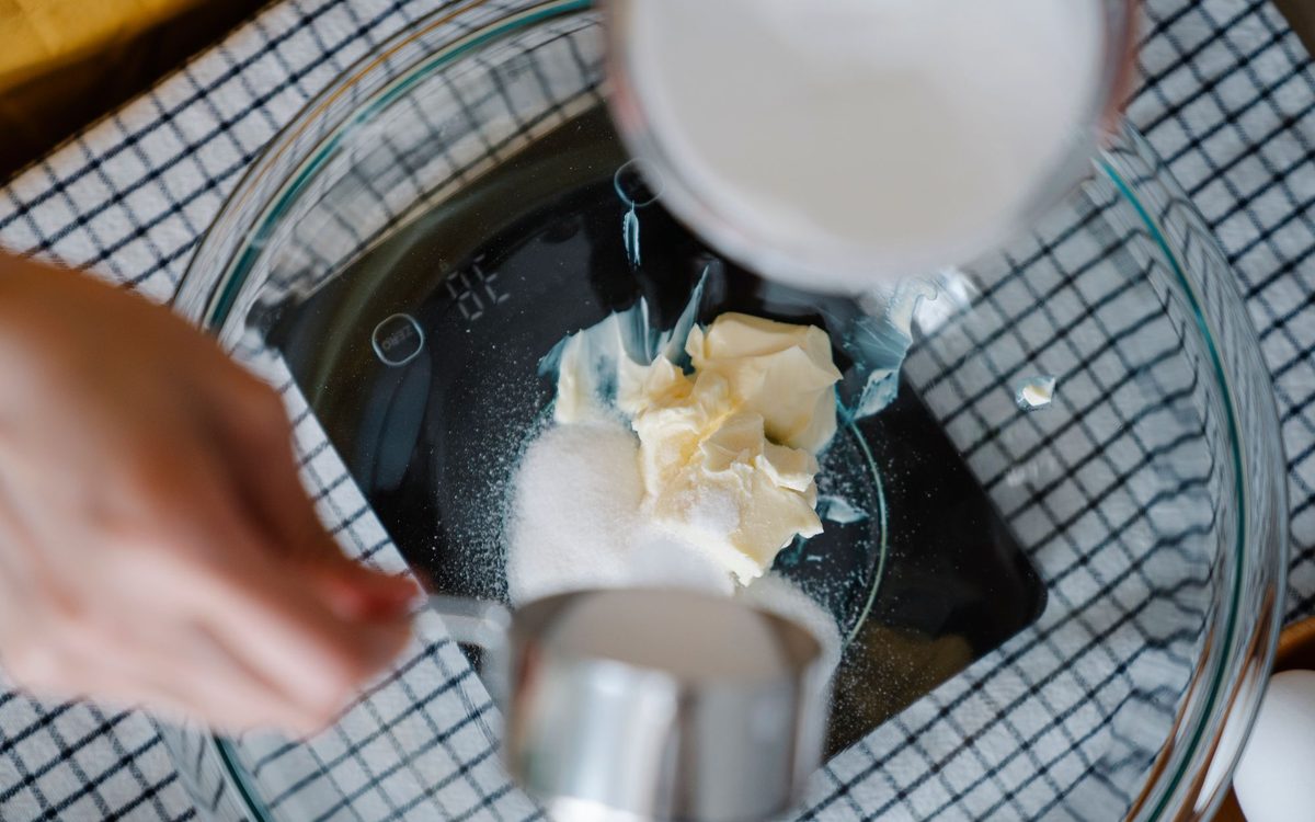 Overhead view of a hand adding sugar from a measuring cup to butter in a glass bowl on a kitchen scale, with a cup of milk above.