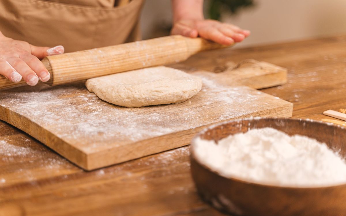 Close-up of hands kneading dough with a rolling pin on a floured wooden board for home baking.