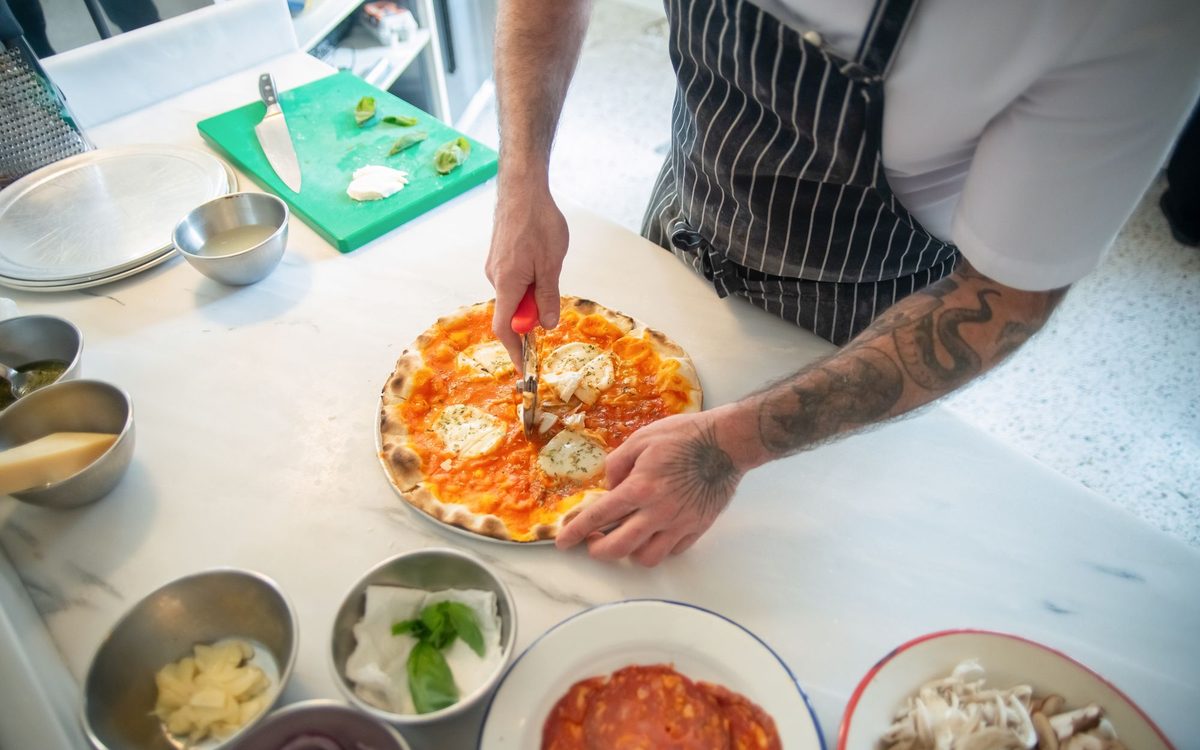 A cook cutting a round pizza with a rotary cutter on a marble counter