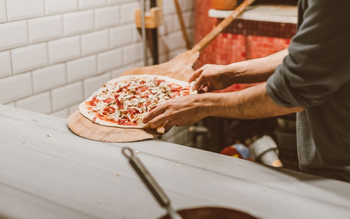 Hands sliding a topped pizza onto a wooden peel, ready to launch into the oven