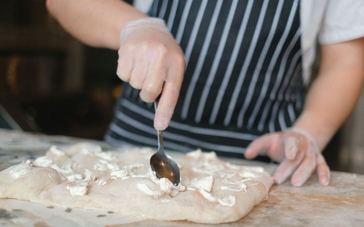 A gloved cook in a striped apron spooning soft cheese onto flattened pizza dough on a counter.