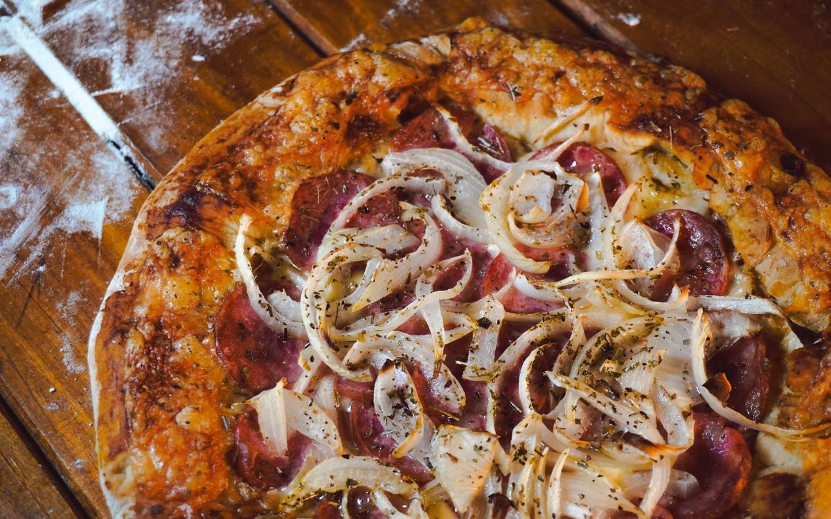A baked pizza topped with salami, sliced onions and herbs, with a deeply browned crust, on a floured wooden table.