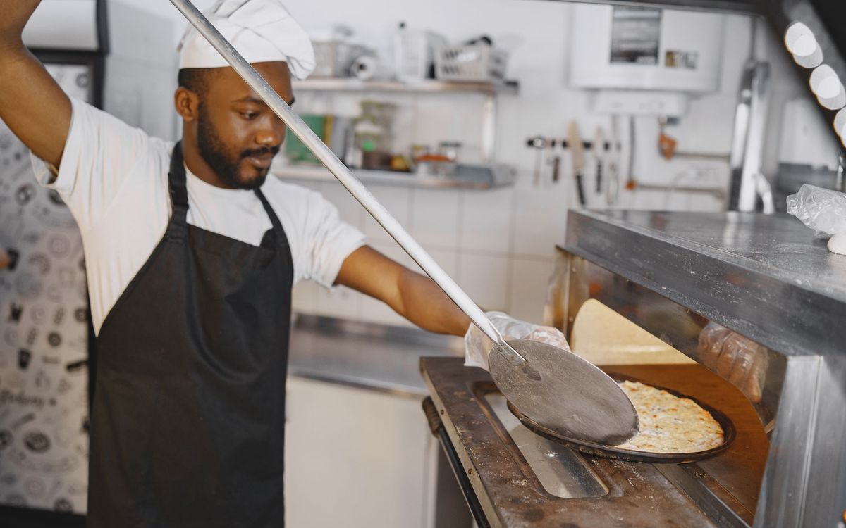 A cook in a white hat and black apron using a long metal peel to slide a cheese pizza into a commercial deck oven.