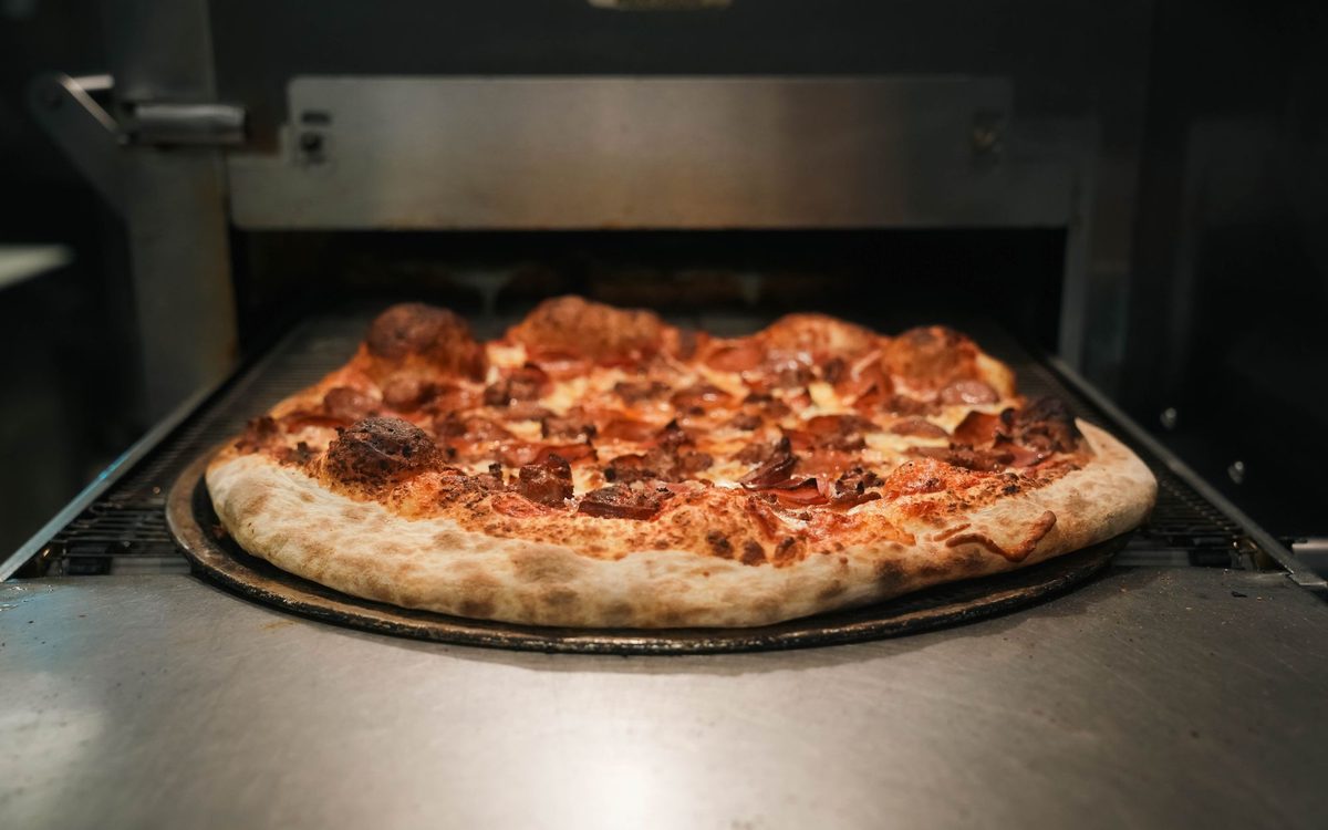 A baked pepperoni and sausage pizza with a browned, blistered crust on a tray at the exit of a commercial conveyor oven.