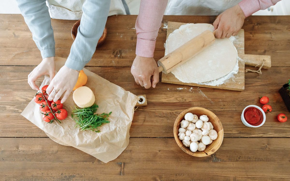 Couple rolling pizza dough and arranging fresh ingredients on wooden table, creating a homemade meal