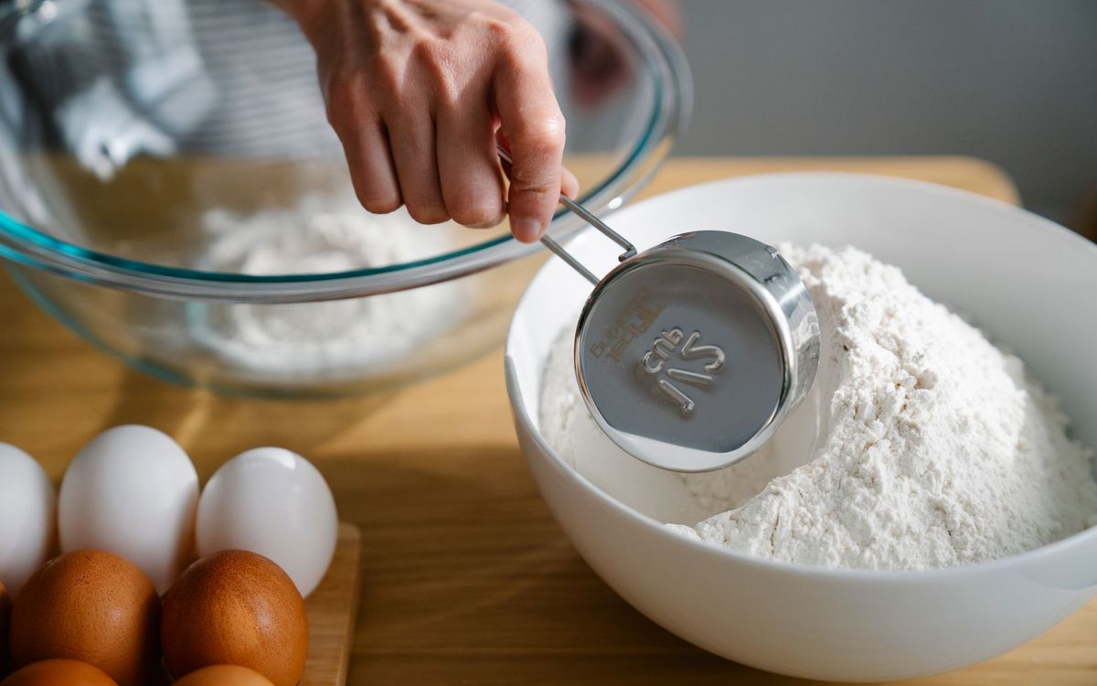 Close-up of a hand measuring flour in a bowl, ready for baking with eggs nearby on the kitchen counter.