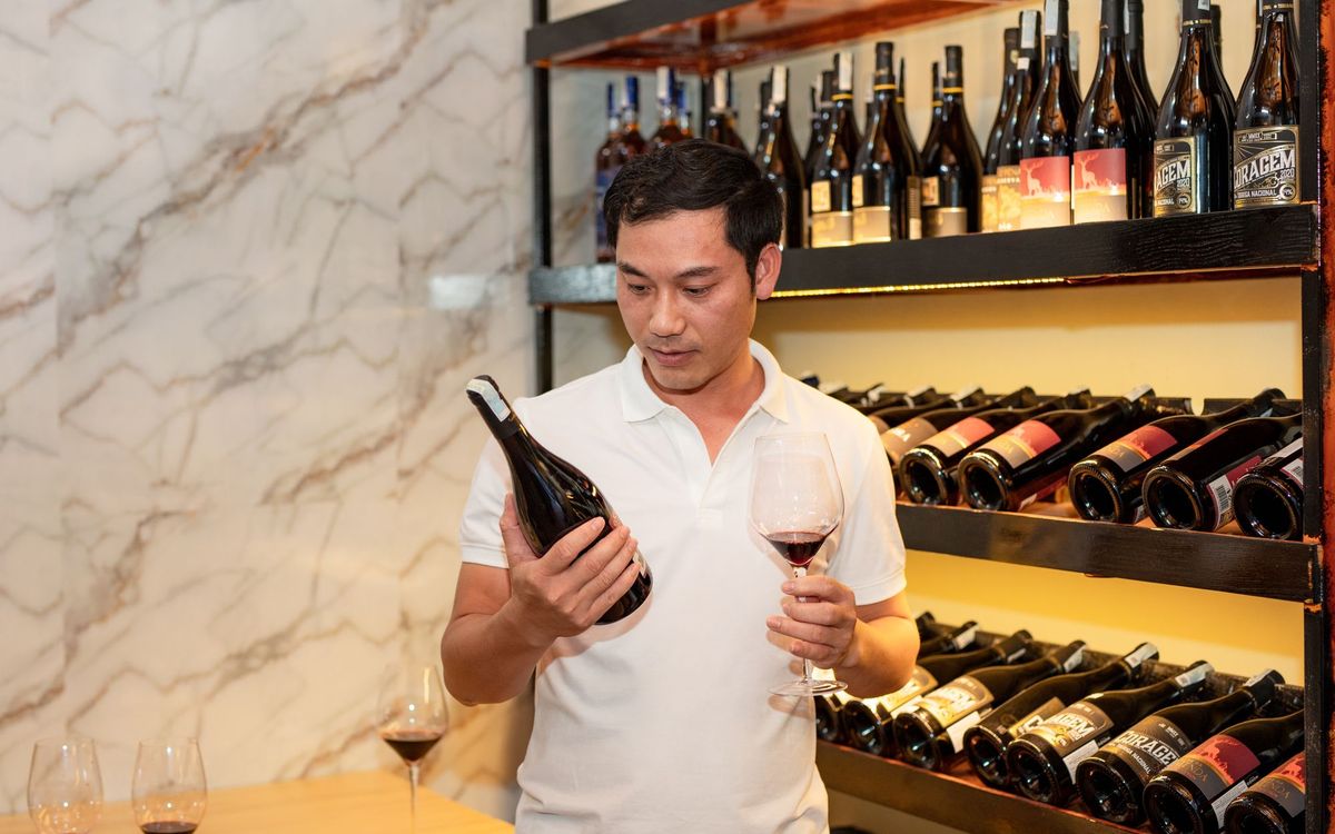 A man in a white polo shirt holds a wine bottle and a glass of red wine beside shelves with bottles stored on their sides.